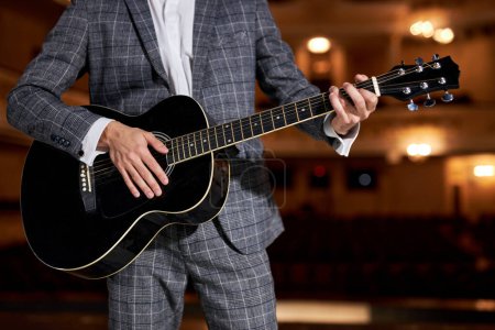 Téléchargez les photos : Cropped european Man in elegant classic suit plays and sings while standing on stage on 12-string acoustic guitar, performance. Lighted background of the theater hall. art, music concept - en image libre de droit