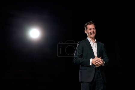 Téléchargez les photos : Man speaking through microphone in dark conference hall, on stage, performance. Person in suit says about marketing and management for successful sales to audience. Professional ideas - en image libre de droit