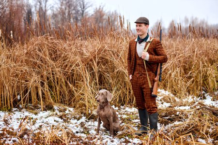 Foto de Male hunter in suit with dog posing looking at side outdoors in nature, with rifle, looking for target wild animal in horizon. handsome guy is ready to hunt, cold weather at autumn season - Imagen libre de derechos