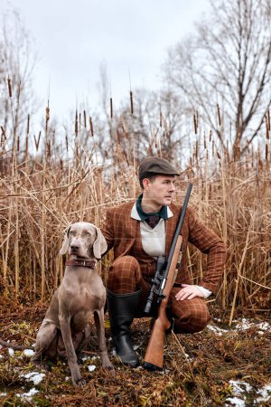 Foto de Serious caucasian hunter man with dog holding shotgun rifle walking in countryside nature, wearing brown trendy stylish suit and hat. young skilled experienced male hunter waiting for prey - Imagen libre de derechos