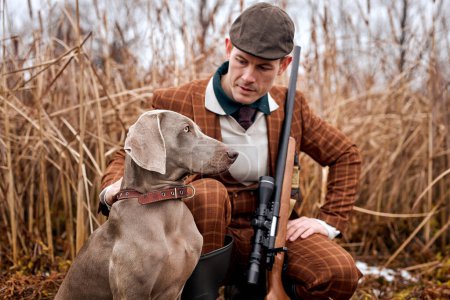 Photo for Hunter with shotgun in suit clothes in autumn forest in search of trophy prey. handsome caucasian man with weapon and hunting dogs tracking down the game. in countryside rural area - Royalty Free Image
