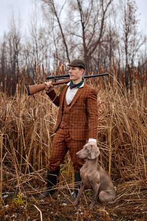 Téléchargez les photos : Shikari with gray hunting dog in autumn forest. Image taken during big game hunting trip. Hunting period, autumn season open. handsome caucasian guy in brown suit outdoors in nature - en image libre de droit