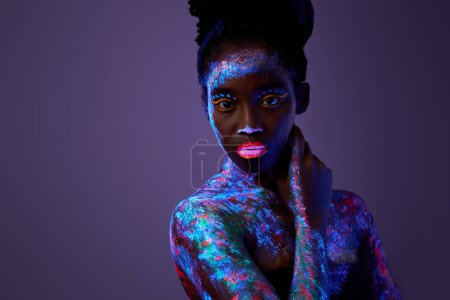 Téléchargez les photos : Charming flawless black woman hold hands on neck wear no clothes isolated on colorful purple neon background, attarctive peaceful lady with fluorescent make-up body art posing at camera - en image libre de droit