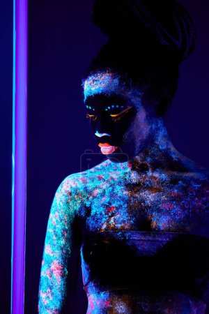 Téléchargez les photos : Femme noire fascinante mince avec l'art du corps au néon avec la poudre fluorescente posant à la caméra seule, en arrière-plan sombre studio. Beauté, Mode, Beauté, Art corporel Concept. Gros plan - en image libre de droit