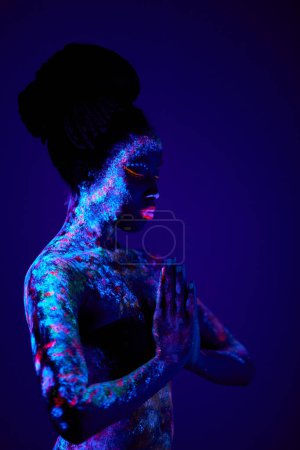 Foto de Young slim black prayer woman with colorful fluorescent prints on skin, beautiful female pray, ask for blessing. prints glows in UV lights. body art, fantasy concept. fantastic, body art concept - Imagen libre de derechos