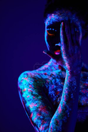 Téléchargez les photos : Mesmerizing black female with fluorescent prints on skin, cosmic paint glowing on neon lights, black background. young calm and peaceful lady losing half of face with hands, close-up - en image libre de droit