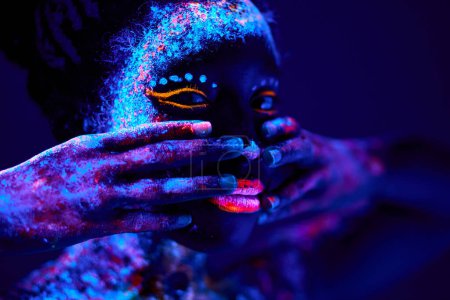 Téléchargez les photos : Beautiful young black female with fluorescent prints on face, unusual prints, body art. neon lights, uv ray, luminescence concept. model touching face, covering. close-up portrait - en image libre de droit