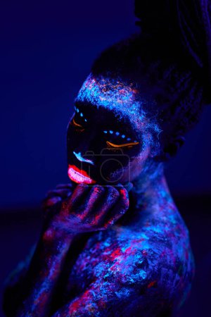 Téléchargez les photos : Femme noire avec art corporel UV luisant dans l'obscurité, impressions colorées sur la peau nue. peintures futuristes fantastiques abstraites, maquillage fluorescent. calme dame consciente posant à la caméra, seul à l'intérieur - en image libre de droit