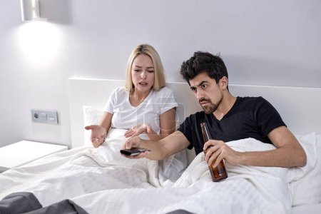 Téléchargez les photos : Blonde woman regretted that she married a mediocre and drunk man, woman screaming arguing at man alcoholic sitting on bed with beer bottles, quarrel. relationships problems concept - en image libre de droit