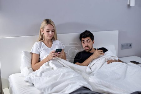 Foto de Infidelity. Cheating Caucasian Woman Texting On Phone With Lover Ignoring Unhappy Jealous Husband Lying In Bed At Home. Female Cheater Having Affair. Jealousy And Unfaithfulness In Relationship. - Imagen libre de derechos