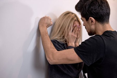 Téléchargez les photos : Family violence. aggressive and nervous bearded guy arguing with crying blonde wife at home, caucasian woman is standing with back against the wall at home, suffering from domestic violence - en image libre de droit