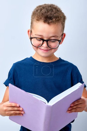 Foto de Smiling kid boy reading fascinating book during school studies against white background, caucasian child in spectacles eyeglasses stand enjoying a new book, school and education concept - Imagen libre de derechos