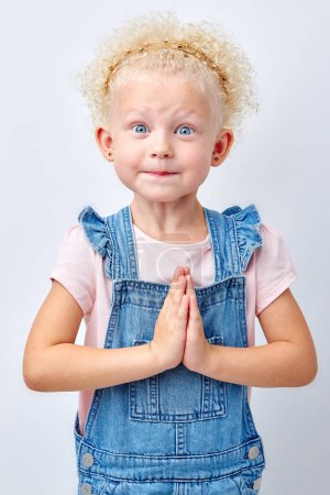 Téléchargez les photos : Portrait of smiling and cute little blonde-haired girl. happy kid lovely keeping hands together isolated over white background, child looking at camera and gesture, pray and dream - en image libre de droit