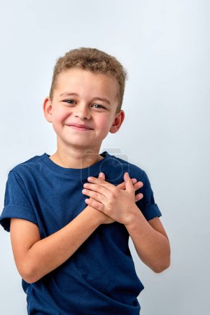 Téléchargez les photos : Close up of cute happy small boy isolated on white studio background hold hands at heart chest feel grateful, smiling little child pray thanking god high powers, faith concept - en image libre de droit
