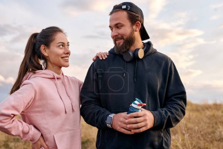 Téléchargez les photos : Outdoor portrait of excited couple in nature countryside relaxing before or after jogging. beautiful woman and man in hoodie laughing, talking. wellbeing, healthy lifestyle, human emotions concept - en image libre de droit