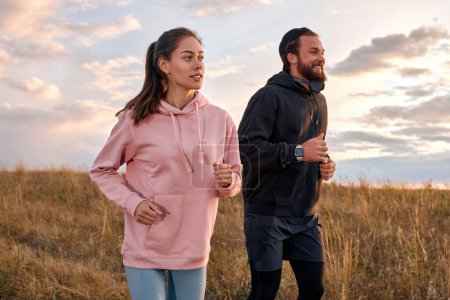 Photo for Couple in sportswear running in field. Active rest on weekends. Traveling in nature. Young caucasian male and woman in sportive outfit engaged in sport, fitness, lead healthy lifestyle. - Royalty Free Image