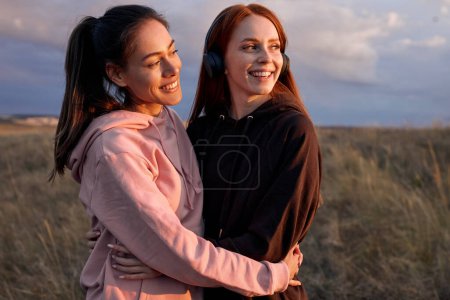 Téléchargez les photos : Portrait Of Positive Caucasian Women Posing At Camera, Having Rest After Jogging, Running Outside Of City, In Field At Sunrise, Laughing, Hugging, Looking At Side. Healthy Lifestyle, Sport Concept - en image libre de droit