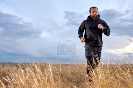 Téléchargez les photos : Handsome Young Caucasian Male Jogging Through the field. Man Wearing Black sportive wear. athlete guy with beard is motivated to be muscular, strong, in perfect physical shape. sport, fitness - en image libre de droit
