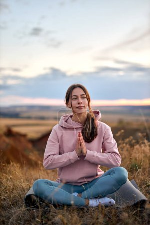 Téléchargez les photos : Pretty caucasian female keep calm, meditating on fitness mat sitting outdoors in nature, keep hands together, engaged in yoga. young lady in sportswear concentrated on mind, enjoying yoga - en image libre de droit