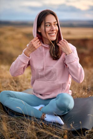 Téléchargez les photos : Happy smiling Adult woman relaxing on mat during yoga exercise, morning routine, sunrise light, outdoor. young female in sportswear stretching at nature open air. sport, healthy lifestyle. - en image libre de droit