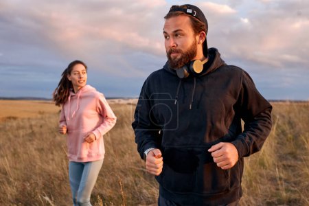 Téléchargez les photos : Couple in sportswear running in field. Active rest on weekends. Traveling in nature. Young caucasian man and woman in sportive outfit engaged in sport, fitness, lead healthy lifestyle. focus on man - en image libre de droit