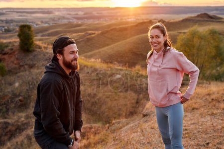Foto de Couple in wonderful field landscape have rest after running, sport training outdoors. Attractive female and confident bearded guy in hoodie looking at side, smiling, having fun, talking - Imagen libre de derechos