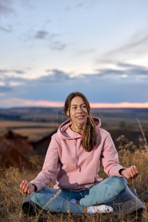 Téléchargez les photos : Smiling Female Keep Calm Sitting On Fitness Mat, Meditating Alone On Fresh Air. Attractive Young Lady In Sportive Leggins And Pink Hoodie relaxing, Having Rest. In Nature In The Morning - en image libre de droit
