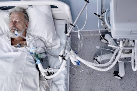 Foto de Sick caucasian senior patient lies on bed in modern hospital , Saline intravenous drip on hand. coronavirus, covid-19, medicine, health concept. Virus and illness protection. top view - Imagen libre de derechos