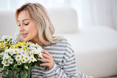 Photo for Charming woman with wildflowers at home with a delicate smile on face. Blonde lady with long hairstyle. charming lady enjoying the smell and beauty of flowers. indoors, in cozy room - Royalty Free Image