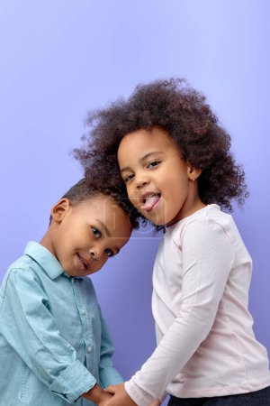 Photo for Happy black children posing at camera and cheerfully smiling over purple background, cute american brother and sister having fun together, showing tongue, wearing casual clothes. familt, children - Royalty Free Image