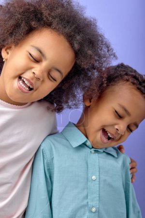 Téléchargez les photos : Two joyful black sister and brother having fun while standing isolated over purple background, portrait of black children in casual clothes laughing, smiling together. human emotions concept - en image libre de droit