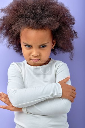 Foto de Naughty child. Disobedience problem. Discipline punishment. Portrait of angry black offended little girl in white with crossed arms isolated on purple copy space background. - Imagen libre de derechos