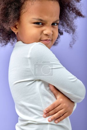 Téléchargez les photos : Angry little black girl showing frustration and disagreement, isolated on purple background, child with curly hair keeping arms folded, in white shirt, posing at camera, offended. portrait - en image libre de droit