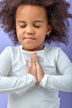 Téléchargez les photos : Cute happy small black girl isolated on purple studio background hold hands together at heart chest feel grateful, smiling little child with eyes closed pray thanking god high powers, faith concept - en image libre de droit
