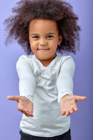Téléchargez les photos : Black cute girl stretches arms for a hug, kind adorable child smiling, isolated on purple studio background. portrait of cheerful curly kid in white shirt posing at camera, charming kid - en image libre de droit