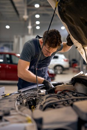 Photo for Young Mid adult mechanic repairing system of car in hood in auto repair shop. view from back on caucasian guy in uniform alone in workshop car service. occupation, garage, vehicle - Royalty Free Image