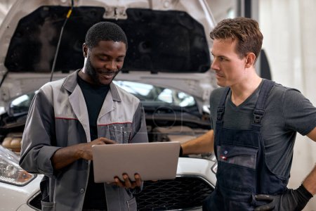 Photo for Handsome diverse mechanics in uniform examining car, using laptop while working in modern auto service. black and caucasian Auto mechanics checking car,service via online insurance system at garage - Royalty Free Image