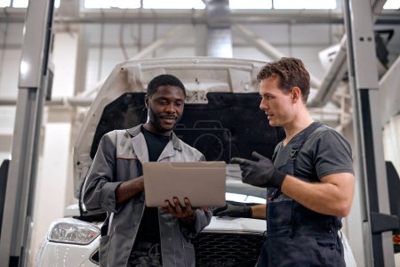 Photo for Black Car Mechanic And Caucasian Repairman Checking Diagnostics Results on Laptop, Computer. Explains an Engine Breakdown to Mechanic. Car Service Employees Inspect Cars Engine Bay. Clean Workshop. - Royalty Free Image