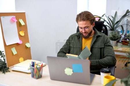 Photo for Smiling handsome office worker man sit reading plan, holding notebook in hands, bearded caucasian male in casual wear concentrated focused on work, generating business ideas. success, office - Royalty Free Image