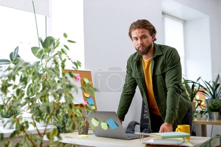 Photo for Bearded young confident guy stand behind office desk with laptop thinking working alone. handsome male in casual wear engaged in office work, plannin strategy, deadlines. at workplace - Royalty Free Image