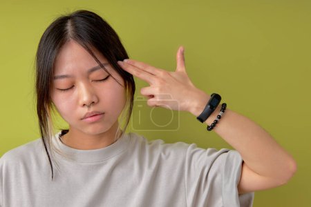 Foto de Serious attractive asian chinese woman in casual clothes holding fingers on temple having thoughtful expression. Female student having clever look isolated over green studio background - Imagen libre de derechos