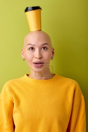 Foto de Frontal portrait of young bald caucasian european woman with cup of coffee on head, stand with surprised facial expression. isolated green studio background. people human emotions concept - Imagen libre de derechos