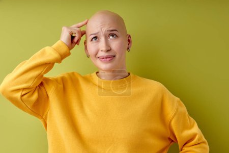 Foto de Studio portrait of pretty bald orhairless woman pondering over something touching head standing on green isolated background. caucasian stressed lady scratching head as seeks, model with alopecia - Imagen libre de derechos