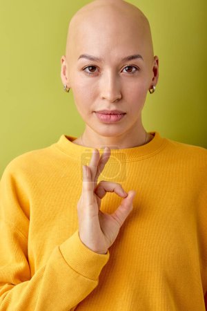 Photo for Confident young bald hairless woman wearing yellow shirt posing isolated on green background studio portrait, looking at camera. Mock up copy space, ad. Showing OK gesture. - Royalty Free Image