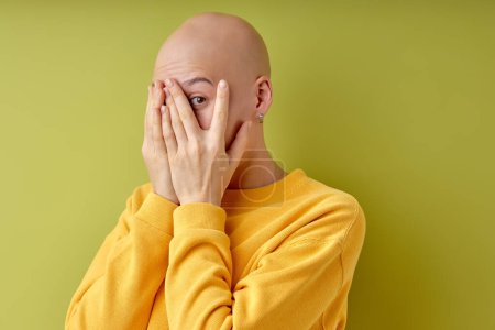 Bald female is closing eyes, frightened and scared about something, isolated on green studio background, copy space. Portrait of emotional lady in casual yellow shirt, allopecia or cancer concept