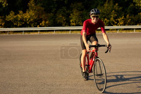 Foto de Handsome sportive male bicyclist riding bike and observing environment in countryside road, confident young caucasian guy in sportive outfit helmet glasses gear engaged in sport, healthy lifestyle - Imagen libre de derechos