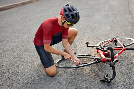 Téléchargez les photos : Athlete man repairs bicycle on road path, outdoor. Hand of cyclist bicyclist examines, fixes modern cycle transmission system. Bike maintenance, top view on guy in t-shirt, shorts annd helmet - en image libre de droit