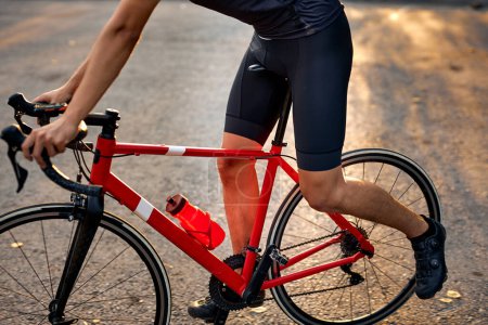 Foto de Close up of sporty young man with strong legs standing on road with red bike. sunset, evening time. Cycling concept. cropped unrecognizable guy in sportive outfit is leading healthy lifestyle - Imagen libre de derechos