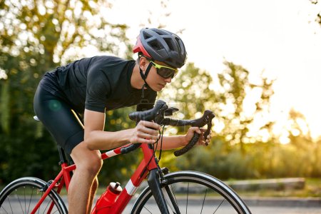 Téléchargez les photos : Strong athlete man in sport clothing and helmet riding professional bike on nature. confident young caucasian sportsman enjoying favorite hobby on paved road. at summer season, sunset - en image libre de droit