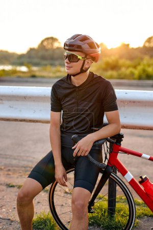 Téléchargez les photos : Athlete sporty cyclist in black helmet, protective glasses and active wear have rest after dynamically riding bicycle. Man after competitions and races on fresh air. outdoors in nature - en image libre de droit